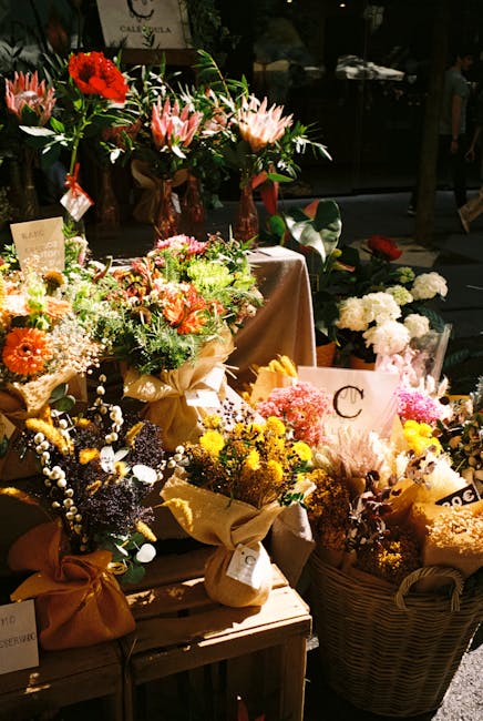 An outdoor display of various floral arrangements featuring vibrant, fresh flowers including pink proteas, red and orange gerbera daisies, and delicate white baby's breath, all arranged in rustic brow